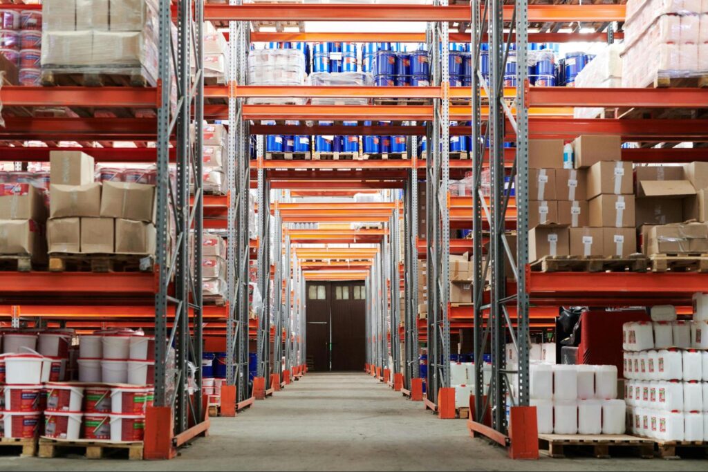 A warehouse aisle with orange shelves, filled with cardboard boxes and white buckets.